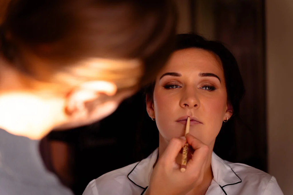 A close-up shot of a bride getting her lipstick applied by a makeup artist while wearing a white pyjama top with dark piping.