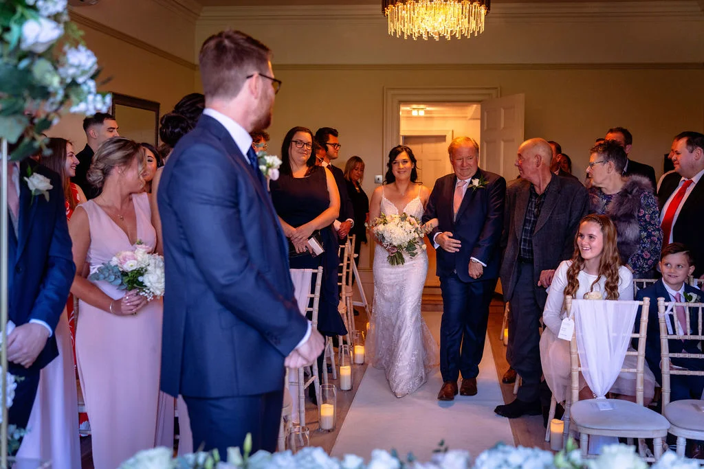 The bride walks down the aisle accompanied by her father, while the groom looks on emotionally from the front during a civil ceremony.