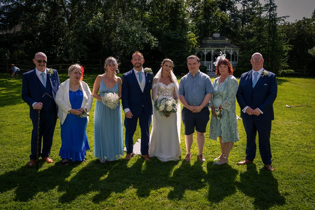 The bride and groom pose outdoors on a sunny lawn alongside family members and wedding party guests in blue suits and dresses.