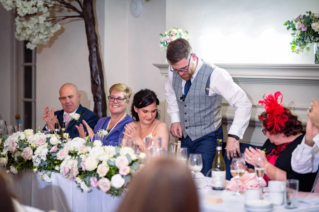 The groom stands at the top table during wedding speeches while the bride and family members applaud and smile.