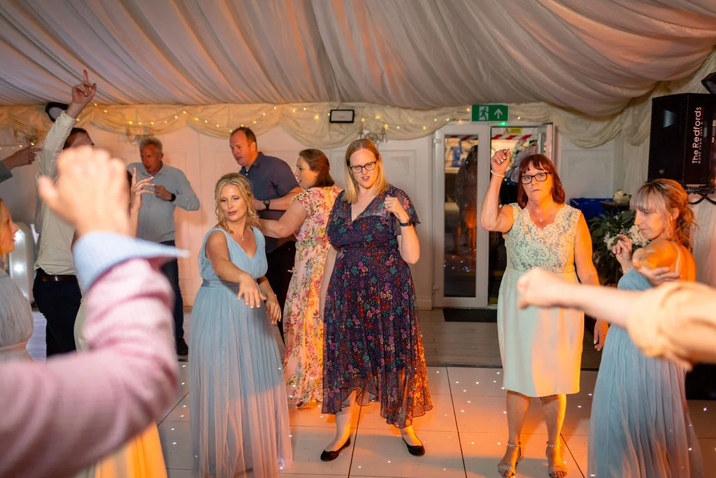 Wedding guests enjoy dancing energetically on an illuminated white dance floor inside a marquee decorated with draped ceiling fabric and fairy lights.