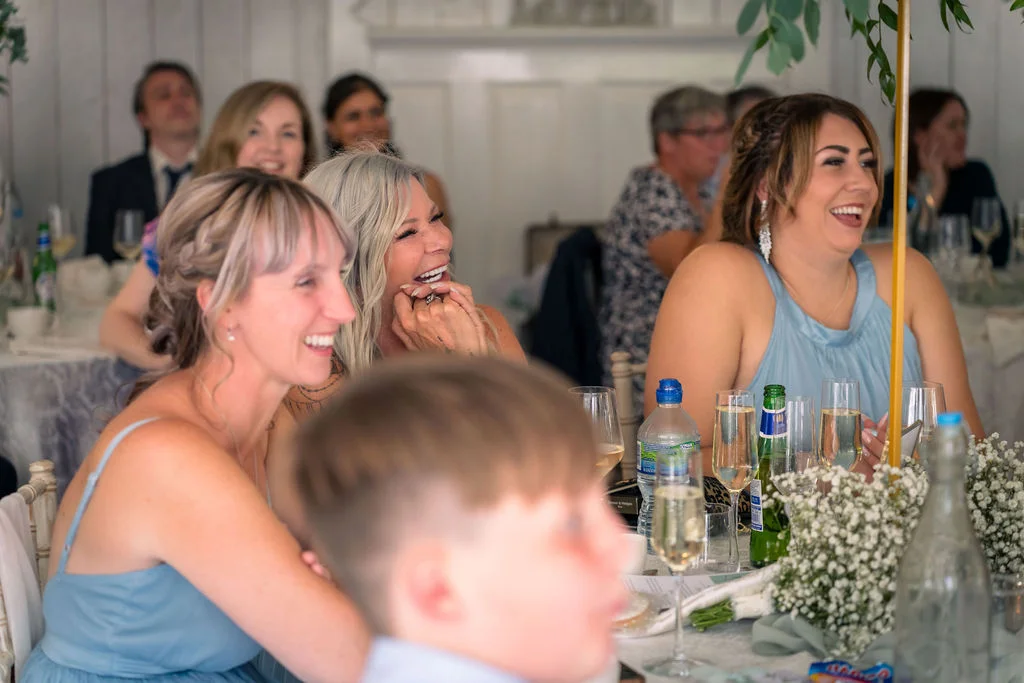 Several wedding guests seated at tables during the reception laugh joyfully together while listening to speeches.