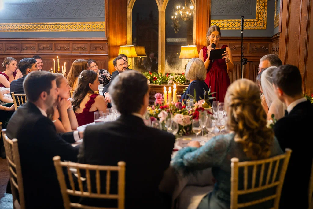 A bridesmaid in a red dress delivers a heartfelt speech using a microphone during the wedding breakfast in a grand wood-panelled dining room.