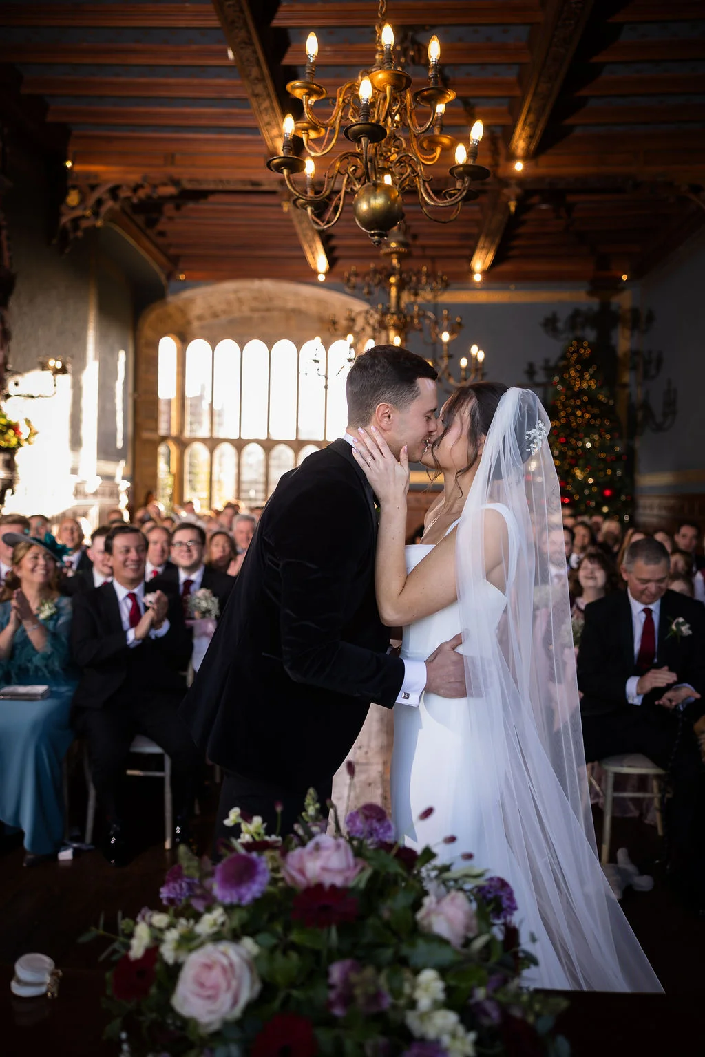 Newlyweds share their first kiss as husband and wife at the front of a grand ceremony room illuminated by a large chandelier and warm window light, with seated guests applauding in the background.