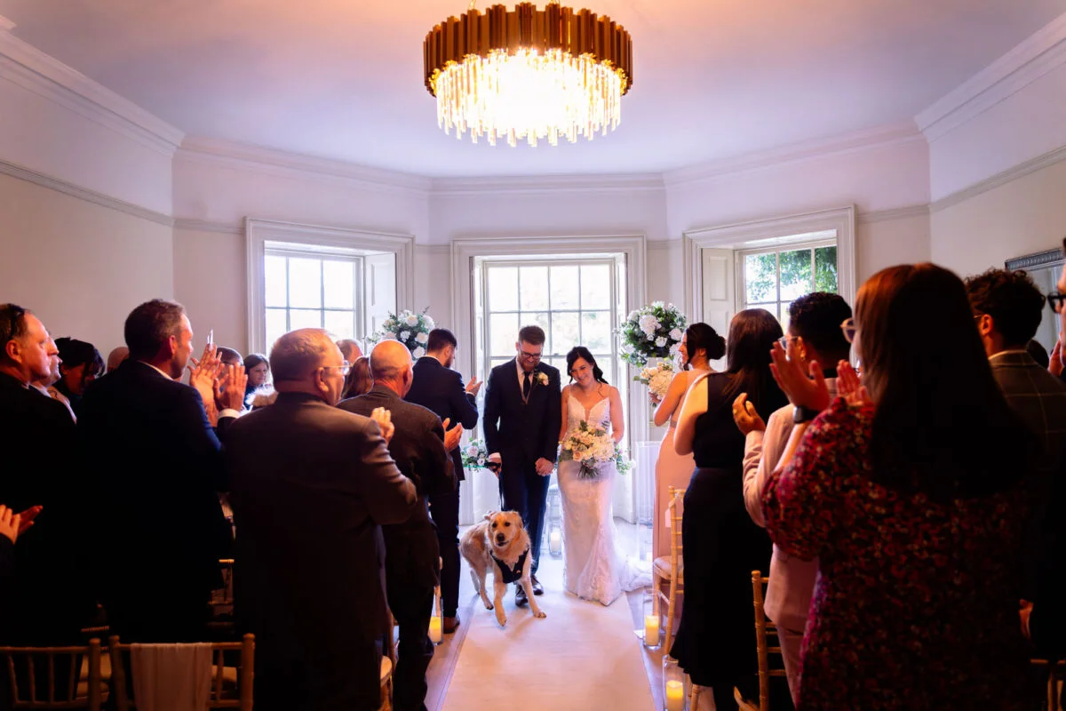 Newlyweds walk down the aisle together with their dog wearing a bow tie, as guests clap and celebrate their ceremony exit.