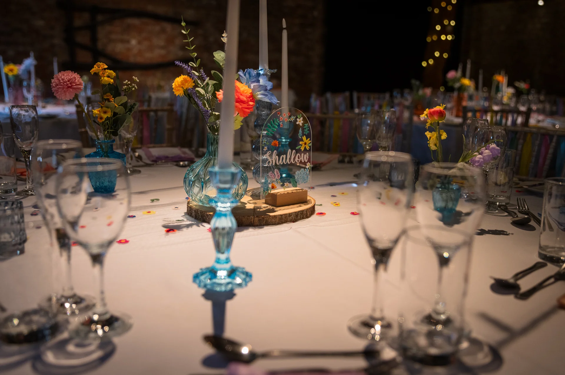 A colourfully styled wedding tablescape features blue glass bud vases filled with bright wildflowers, taper candles in blue candlesticks, and an acrylic table number on a wood slice.
