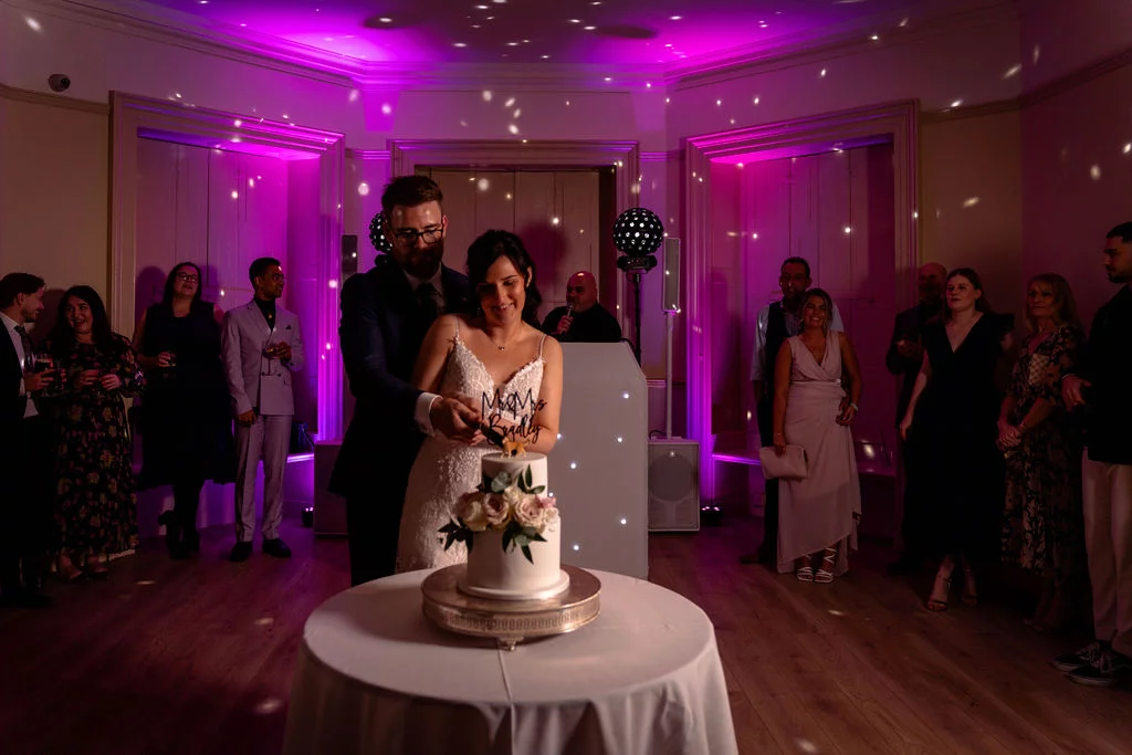 The newly married couple cut their tiered wedding cake together in a ballroom illuminated by vibrant pink uplighting and disco ball reflections while guests watch.