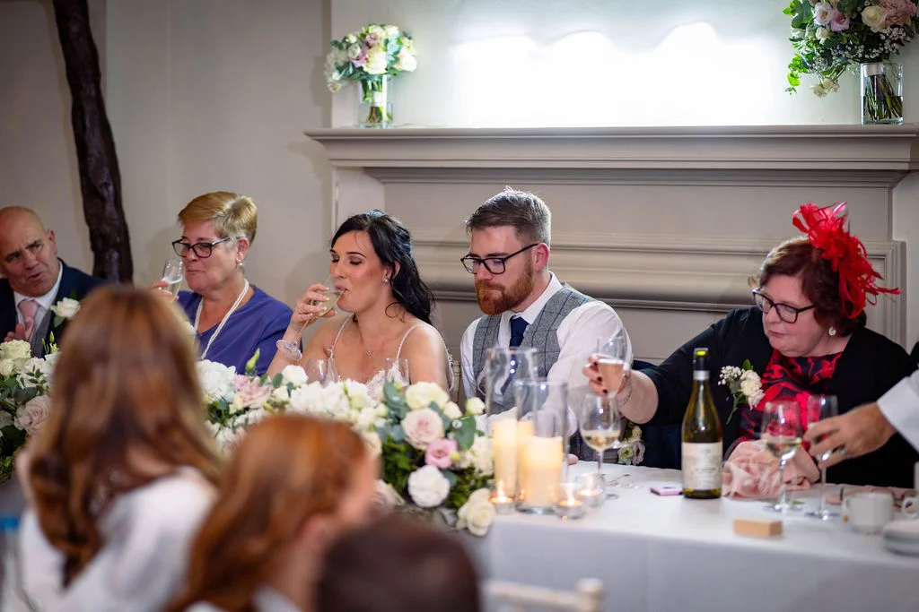 The bride and groom sit at the top table during wedding speeches, surrounded by family members raising glasses of champagne near a fireplace decorated with floral arrangements and candles.