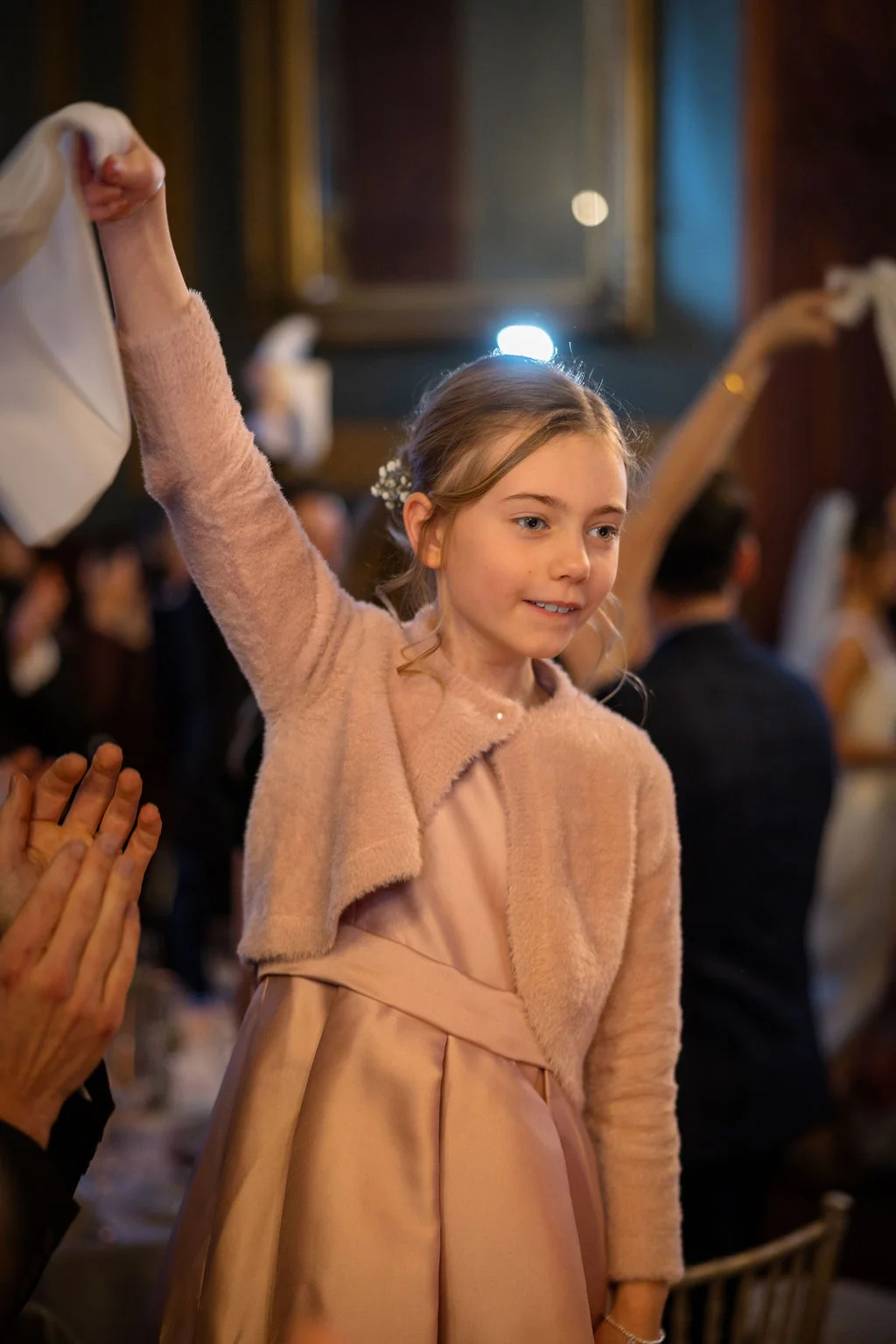 A young flower girl wearing a pink dress and matching cardigan cheerfully waves a white napkin in the air during the wedding reception.
