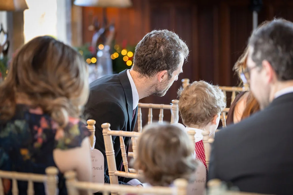 A candid view over the shoulders of wedding guests sitting in wooden chairs inside a warm, wood-panelled venue during a ceremony.
