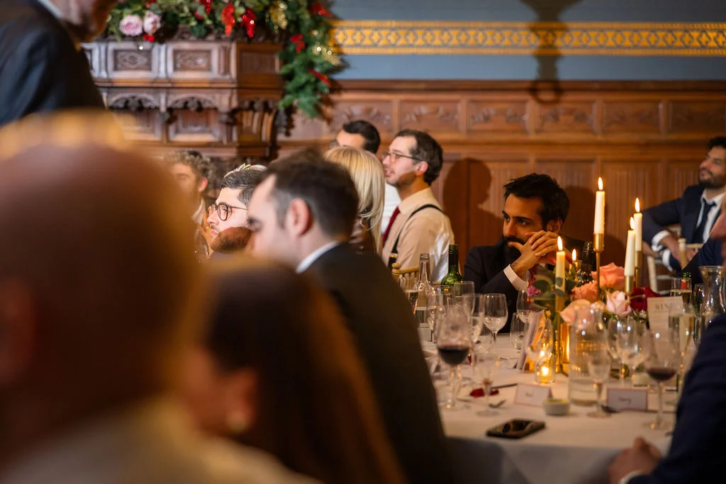 Wedding guests attentively listen to speeches during a candlelit banquet in a historic panelled room with a traditional fireplace.