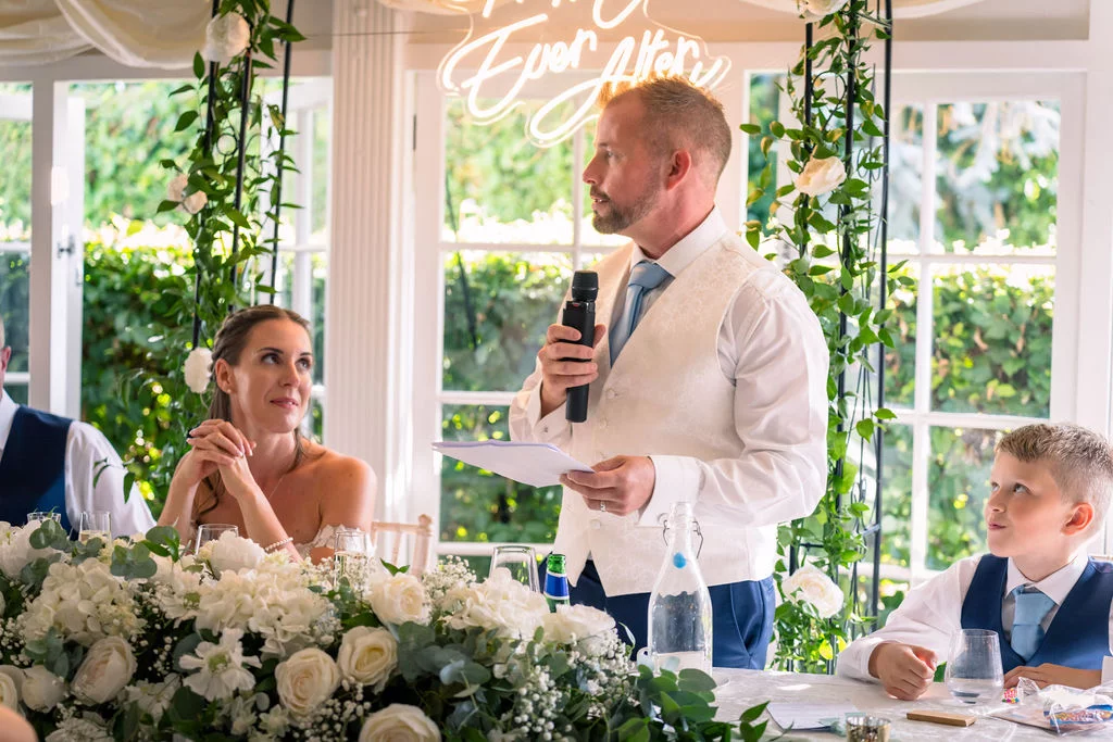 A groom delivers his wedding speech at the top table, holding a microphone and notes, while the bride and a young guest look on attentively against a backdrop of white florals and a neon sign.