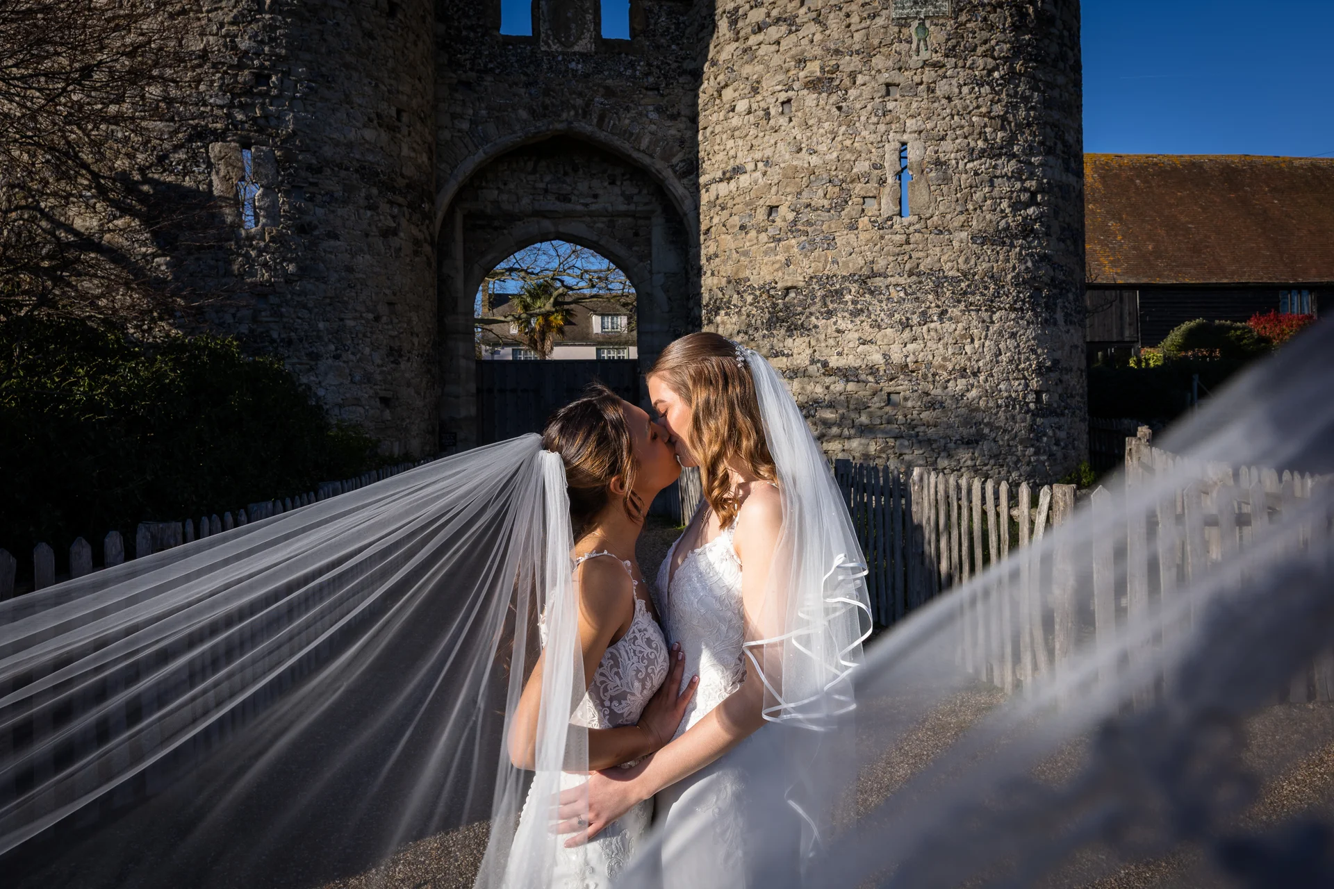 Two brides share a romantic kiss outside a historic stone castle entrance, framed dramatically by a sweeping white veil.