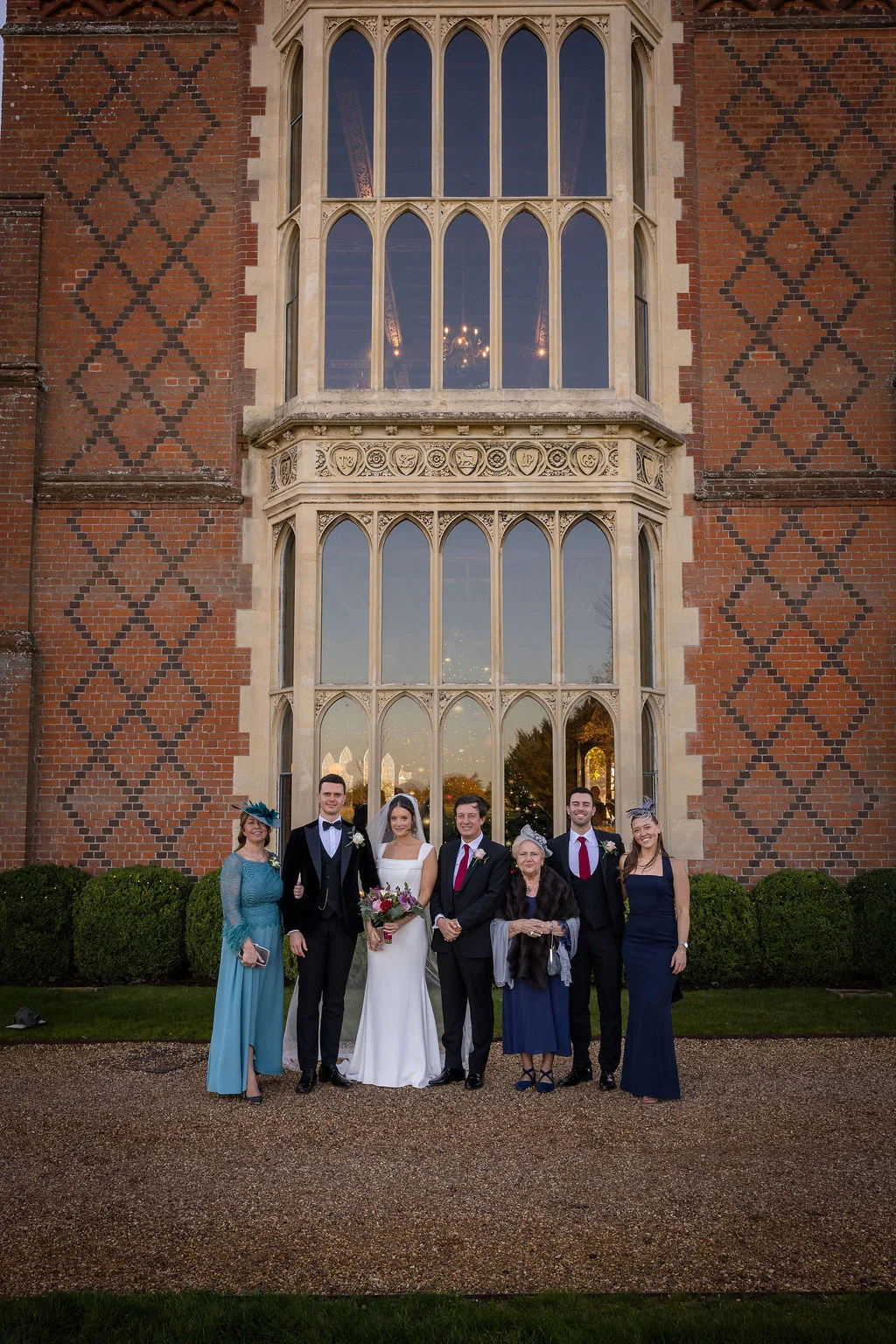 The bride and groom pose for a formal group portrait with family members outside the grand brick facade and large arched windows of a stately home.