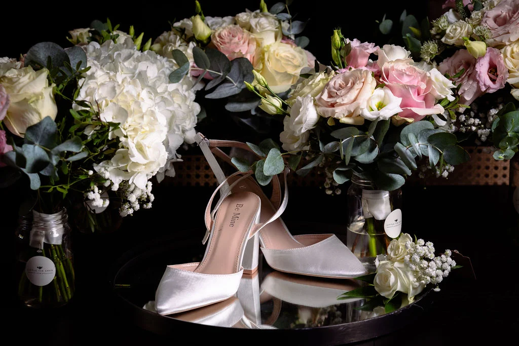 A pair of satin bridal block heels rests on a round mirror surrounded by lush floral arrangements of roses, hydrangeas, and eucalyptus against a dark background.