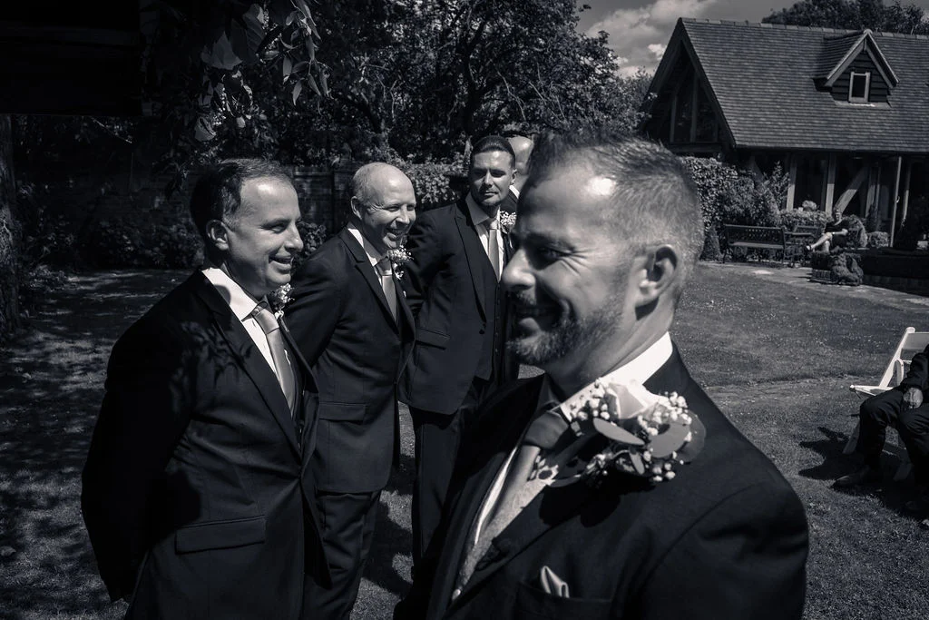 A candid black and white shot of smiling groomsmen gathered outdoors in a sunny garden setting wearing formal suits with buttonholes.
