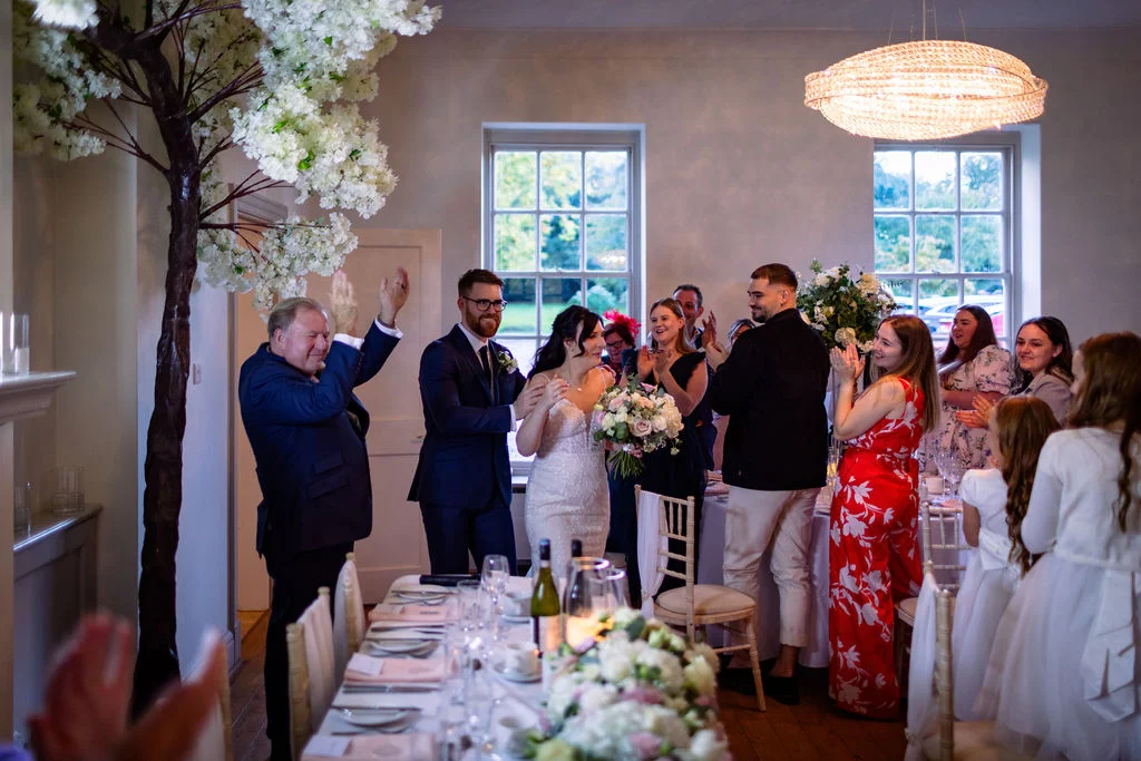 Newlyweds enter the wedding reception to cheering guests and applause, standing near a decorated top table inside a bright, elegant venue.