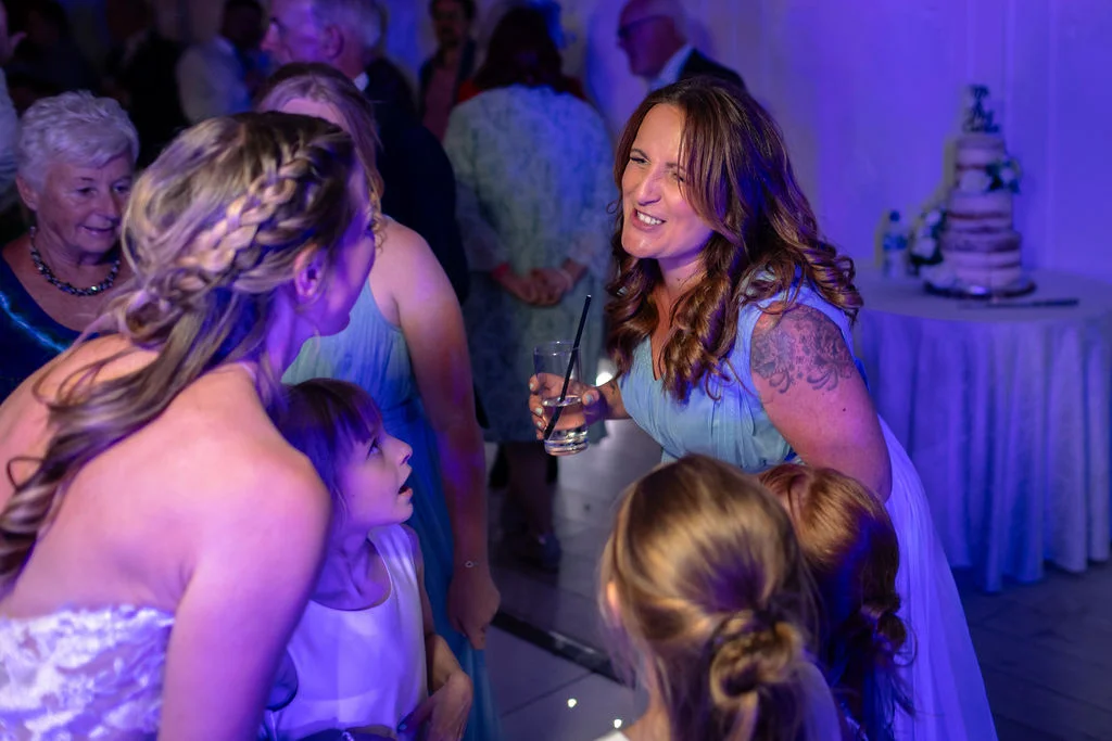 A joyful wedding guest in a light blue dress laughs and chats with the bride during the evening reception under blue lighting.