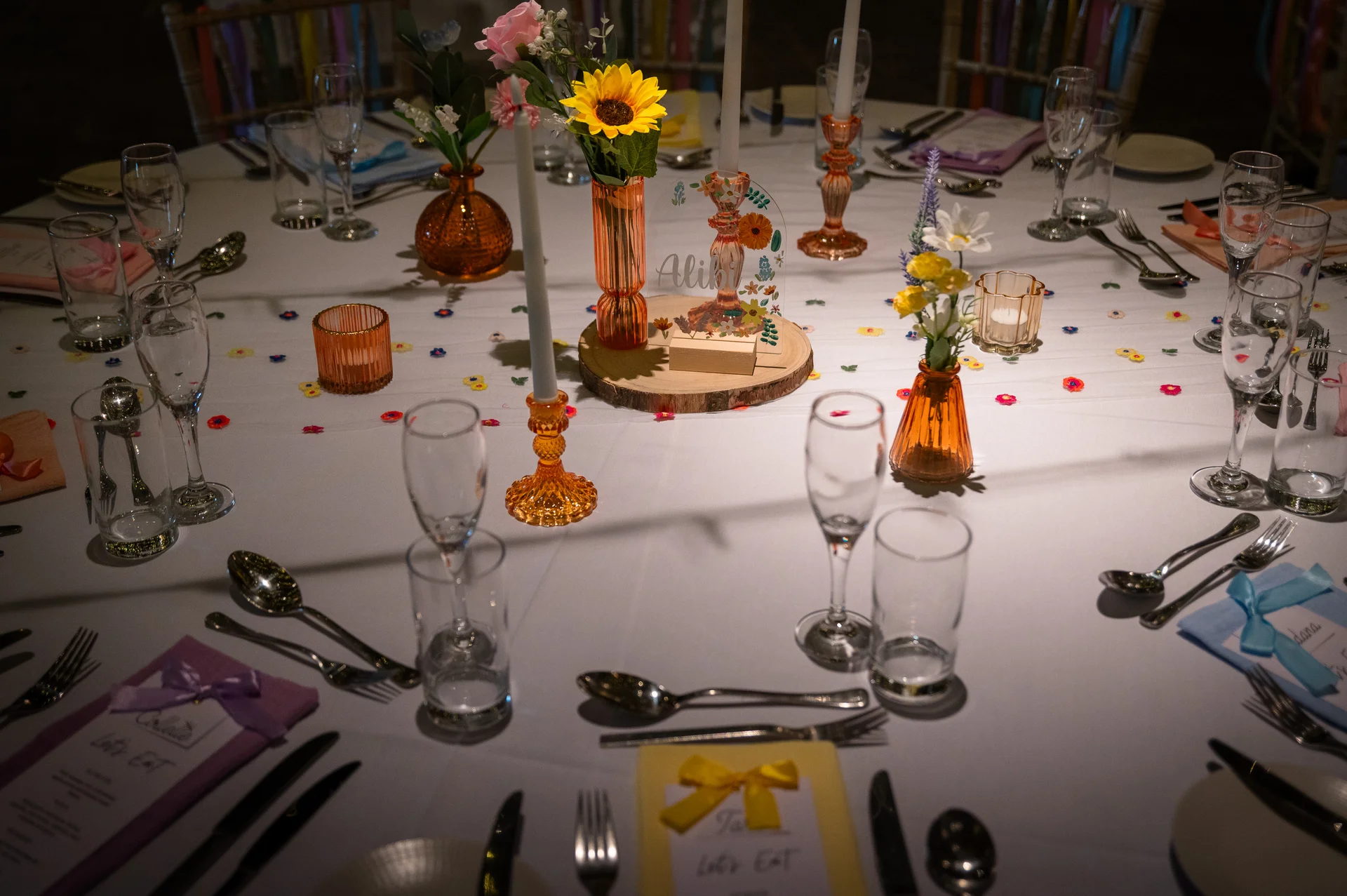 A brightly decorated round wedding table features amber glass vases filled with sunflowers, colourful taper candles, and whimsical acrylic table stationery.