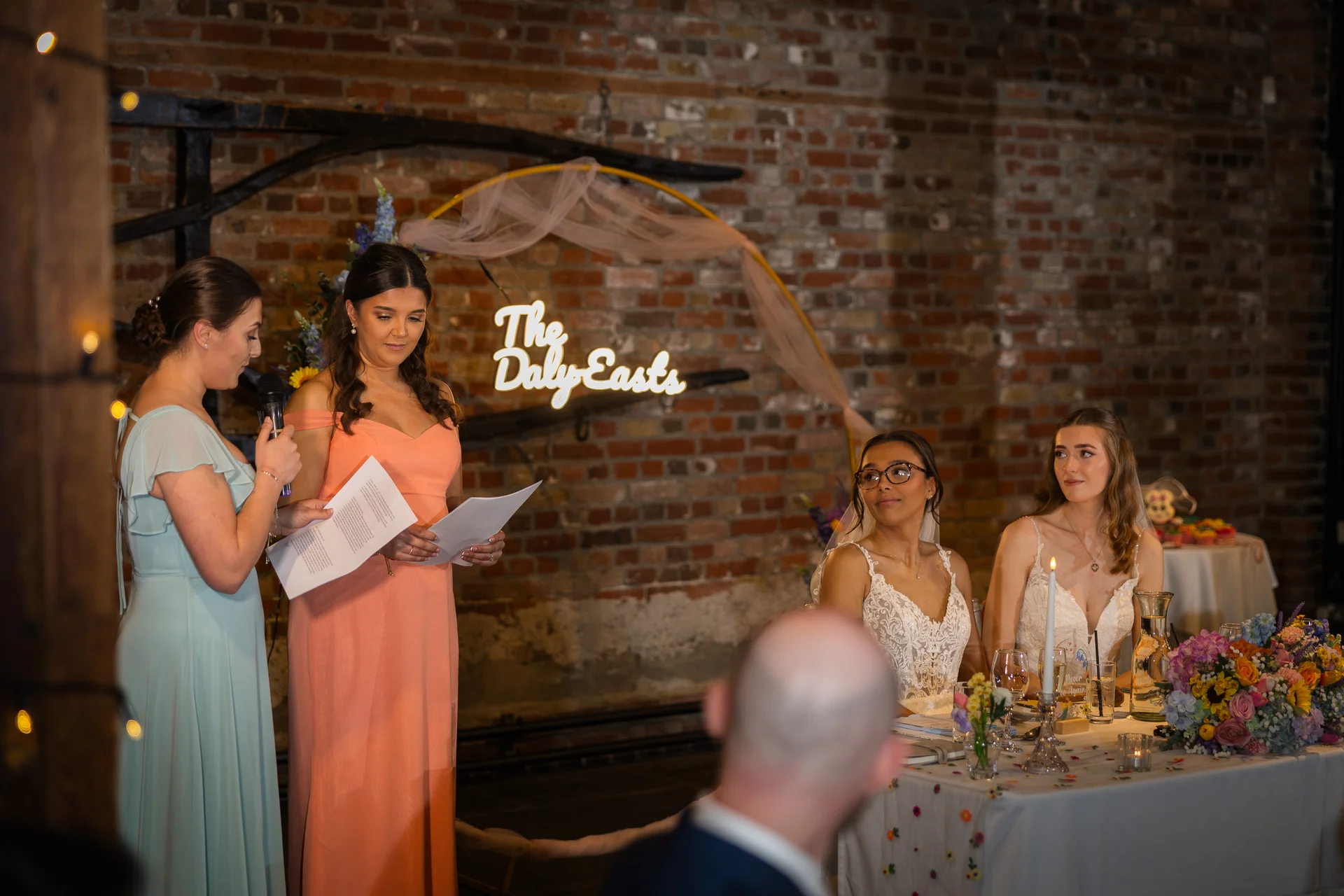 A bridesmaid delivers a speech holding notes at a wedding reception set against an exposed brick wall with an illuminated neon sign.