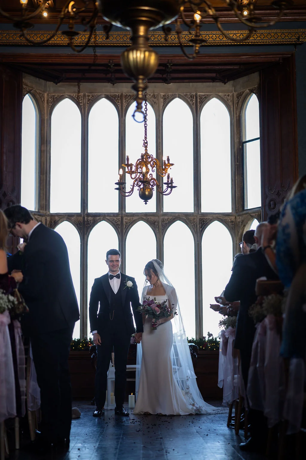 A newly married bride and groom stand together holding hands at the end of the aisle inside a grand historic venue with dramatic arched windows and chandeliers.