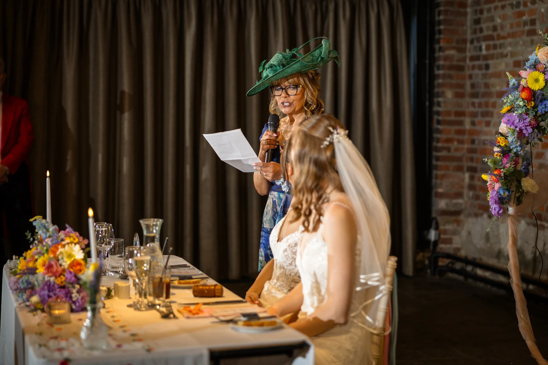 A mother of the bride wearing a green fascinator and glasses delivers a speech holding notes and a microphone at the top table, while the bride listens attentively.