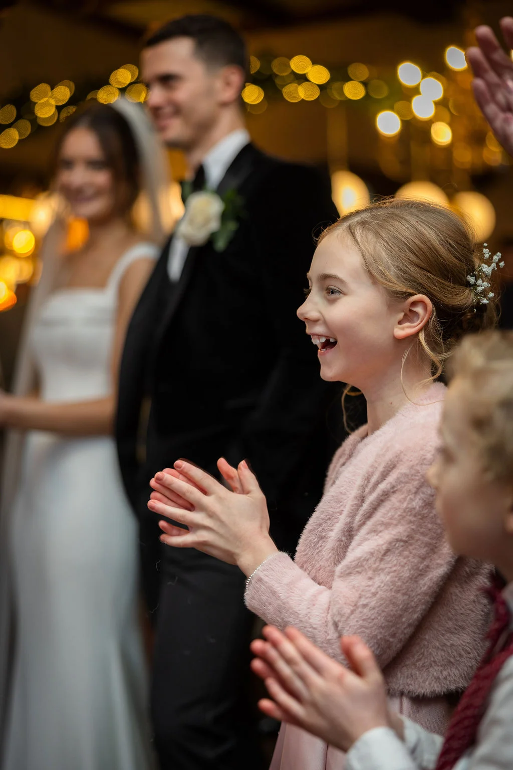 A young flower girl laughs and claps with pure joy as the bride and groom look on happily in the softly lit background.