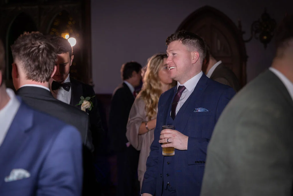 A smiling guest in a navy three-piece suit holds a drink while mingling with others during a wedding reception.