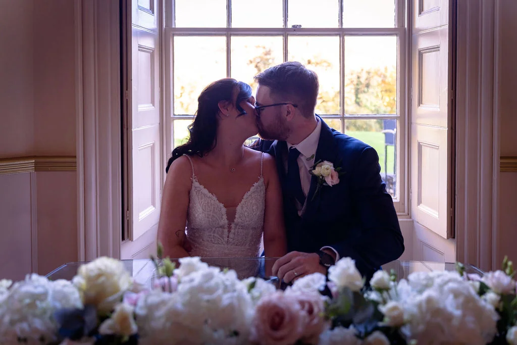 A newlywed bride and groom share a tender kiss framed by a bright window, with a lush floral arrangement in the foreground.