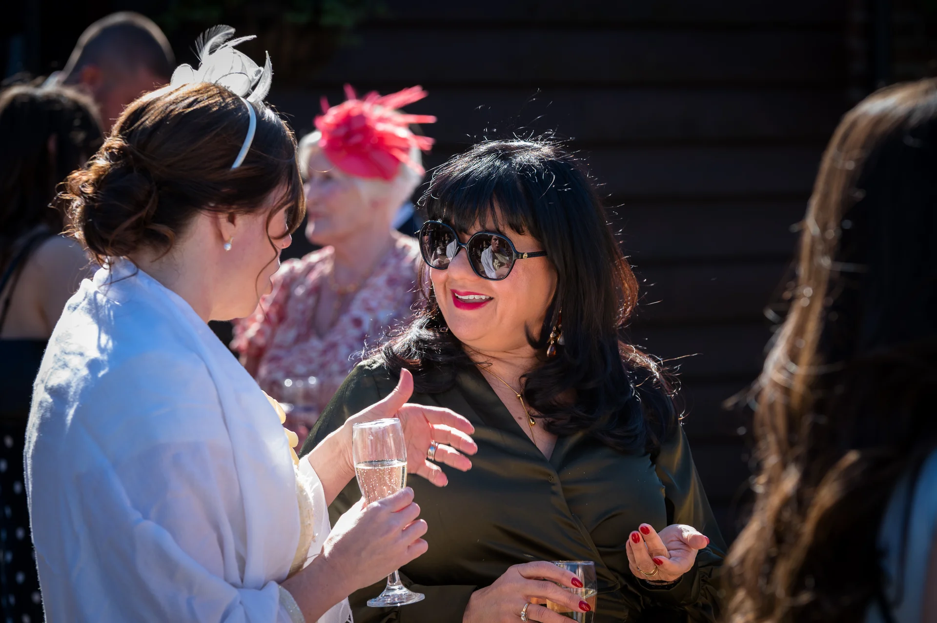 Wedding guests mingle outdoors in the bright sunshine, enjoying conversation and holding glasses of champagne during the drinks reception.