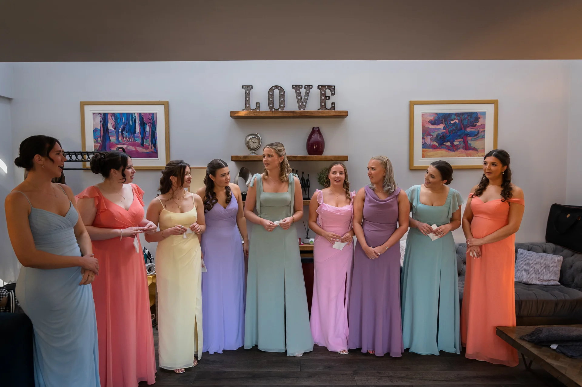 A group of nine bridesmaids in colourful, pastel-toned dresses stand together indoors, chatting and holding small notes while getting ready.