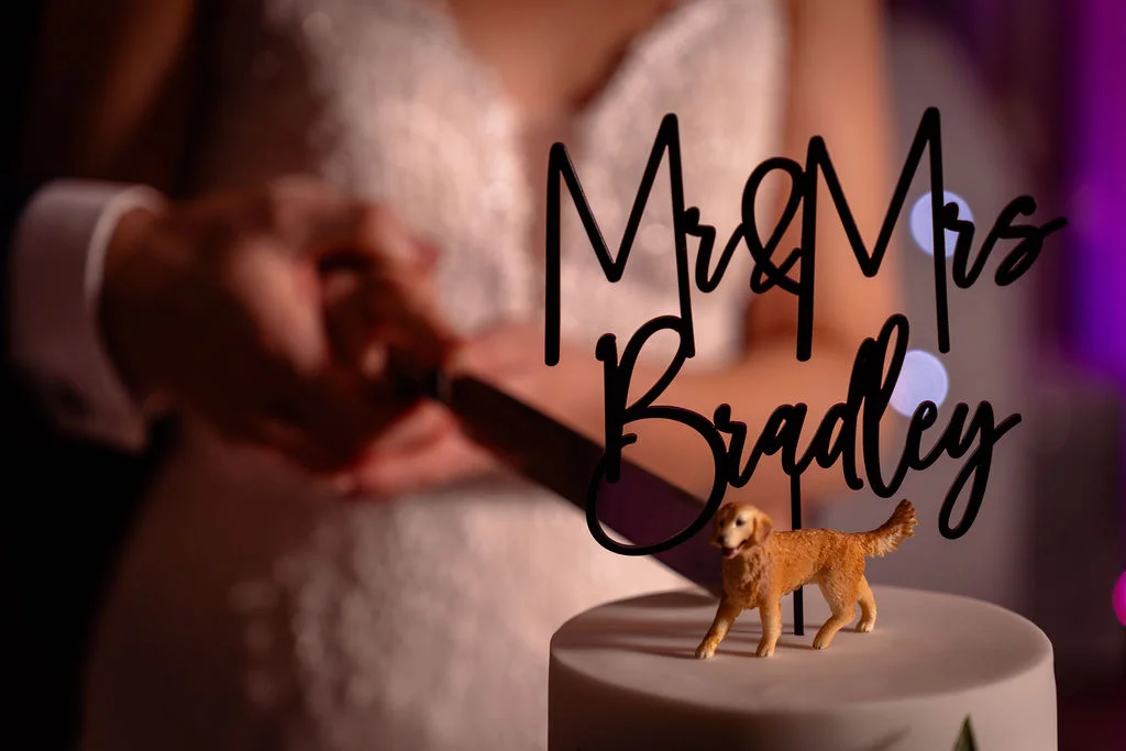 A newlywed couple hold a knife together to cut their wedding cake, featuring a custom topper with a small golden retriever figurine.
