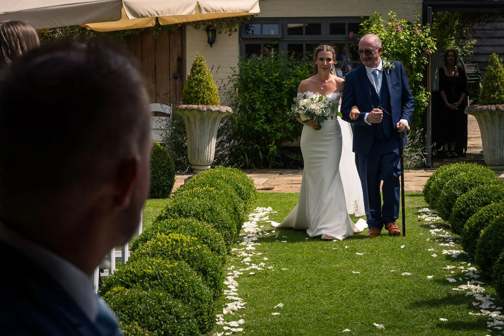 A bride walking down an outdoor garden aisle lined with topiary and white petals, escorted by her father in a navy suit, as the groom looks on from the foreground.