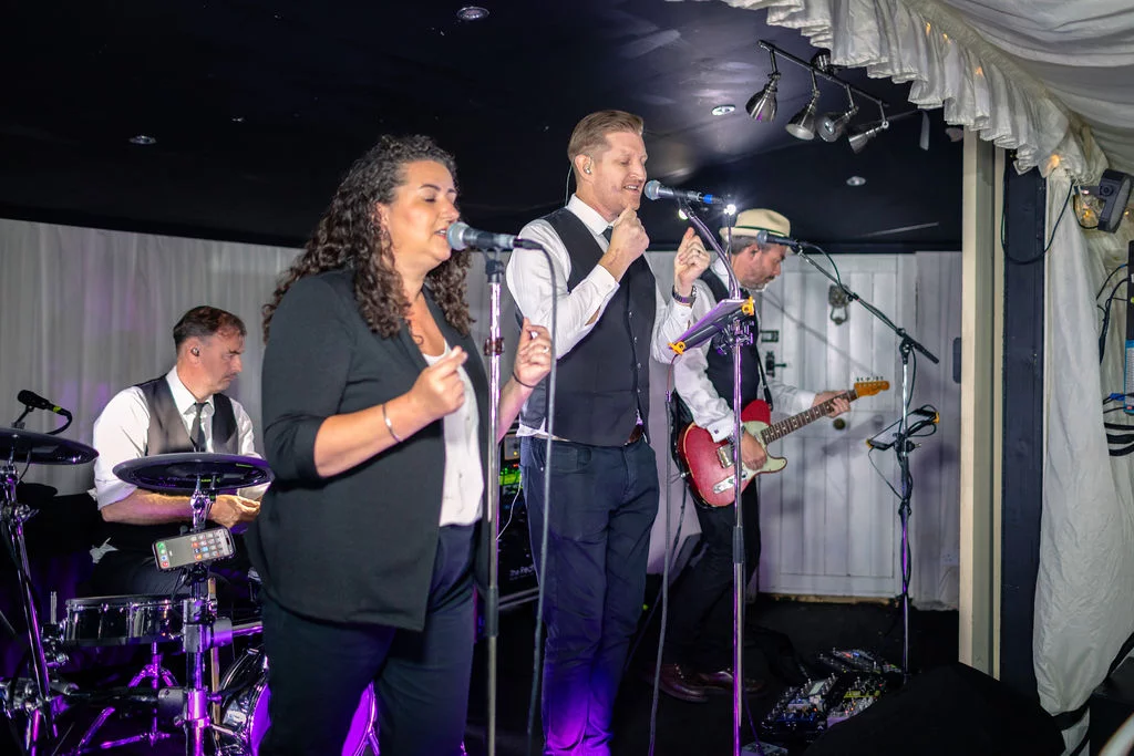 A live wedding band performing on stage in a marquee during the evening reception, with a female singer, male lead vocalist, drummer, and guitarist entertaining the guests.