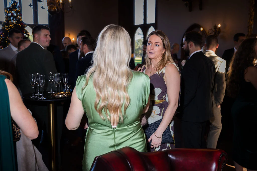 Wedding guests mingle during a lively drinks reception inside a stately home with tall windows and a festive tree in the background.