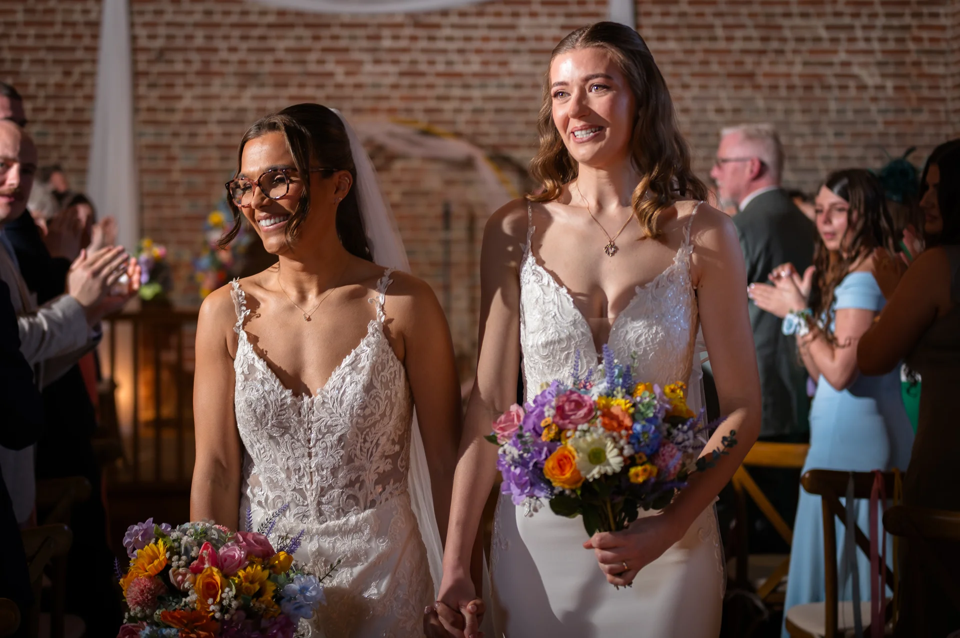 Two smiling brides walk hand in hand down the aisle holding colourful bouquets, surrounded by cheering guests in a rustic brick venue.