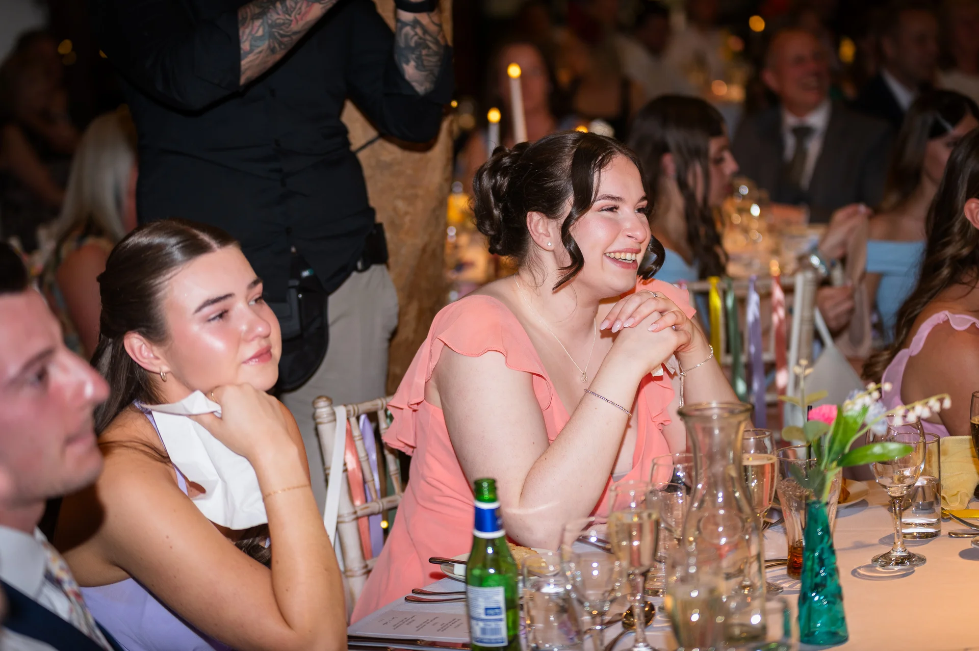 Wedding guests seated at a banquet table laugh and tear up during emotional speeches at the wedding reception.