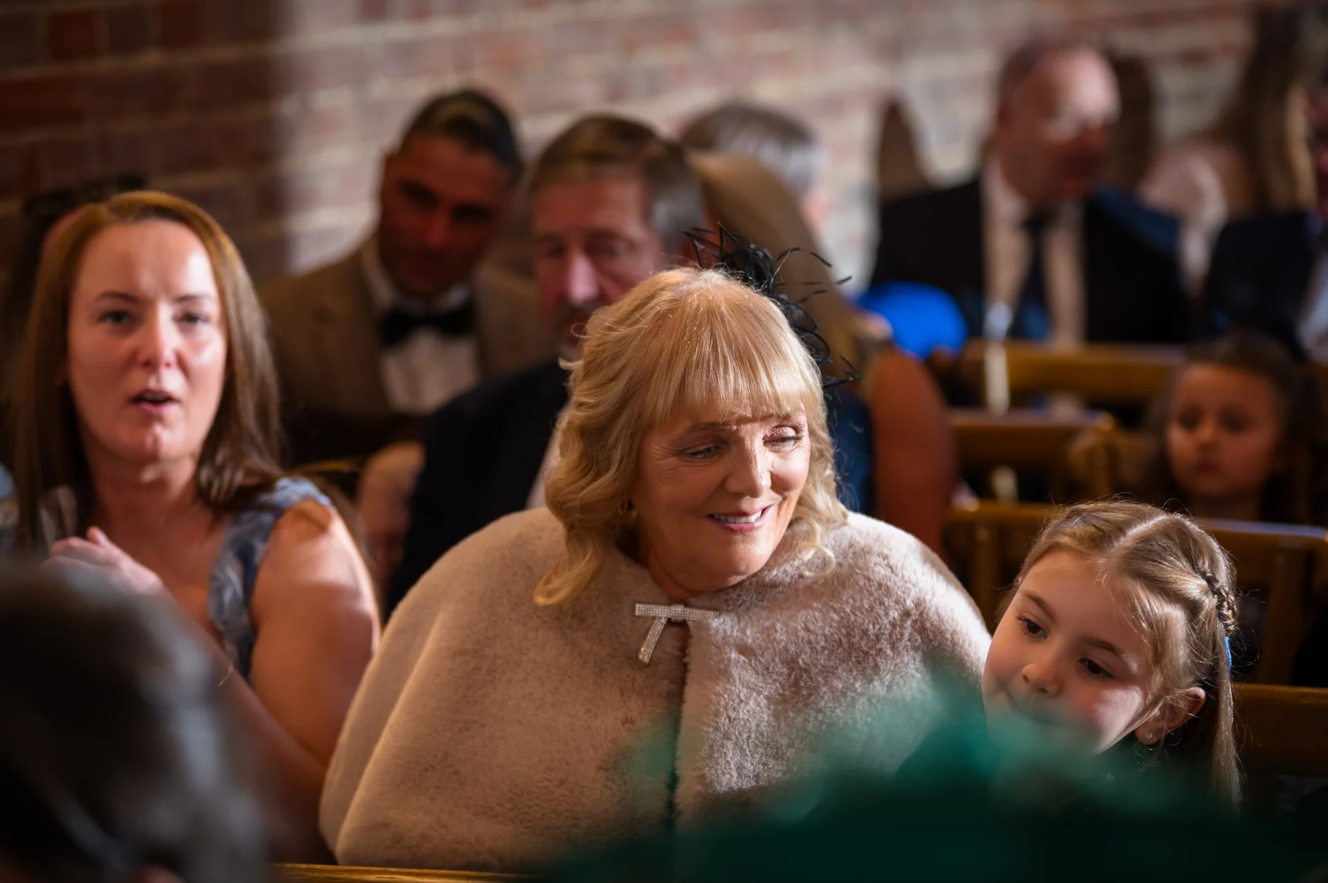 A smiling older woman wearing a faux fur cape and fascinator sits among wedding guests in a venue featuring exposed brick walls.