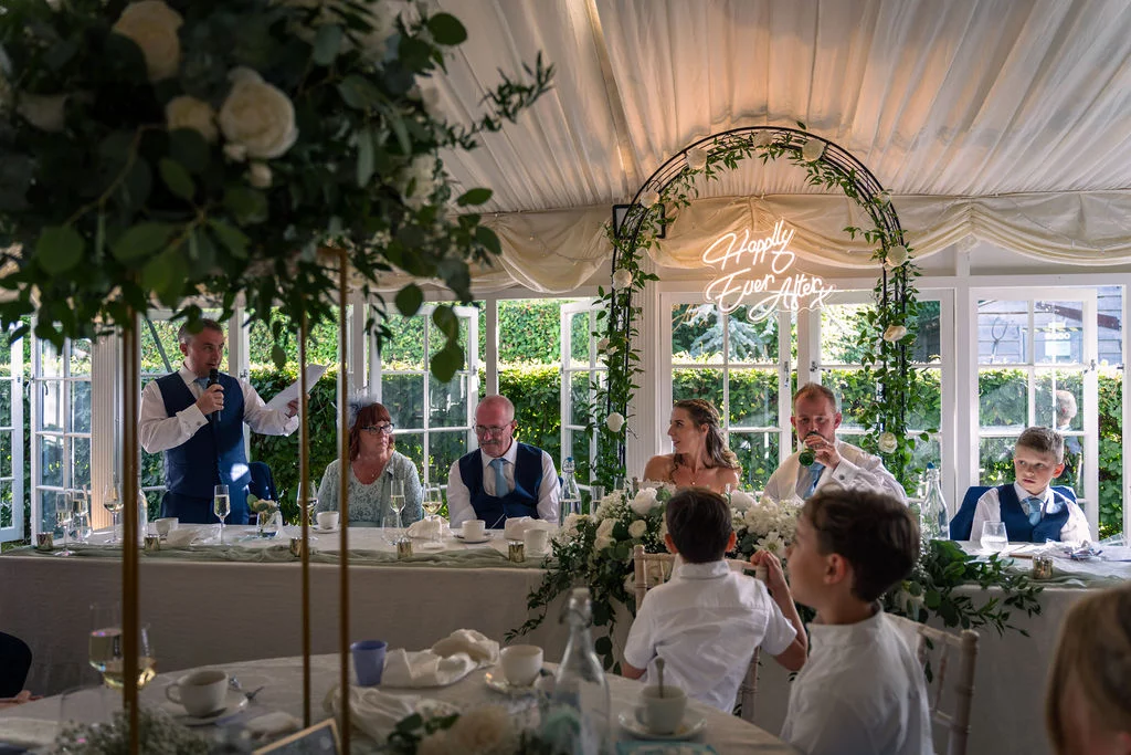 A groom delivers a speech at the top table inside a white draped marquee decorated with a floral arch and a glowing neon sign.
