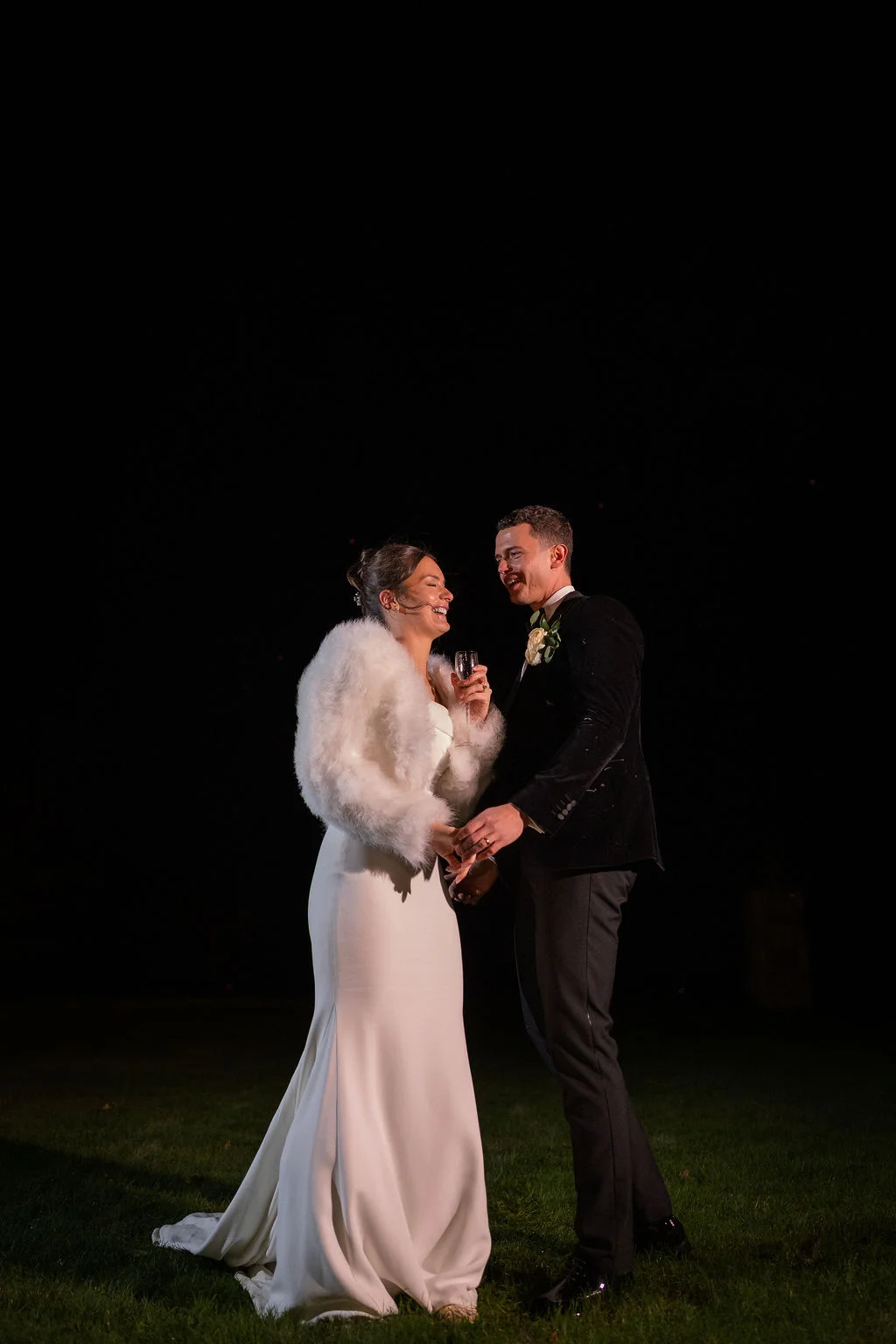 A newlywed couple laughing together outdoors at night, with the bride wearing a white gown and faux fur stole holding a champagne glass while the groom wears a black tuxedo.