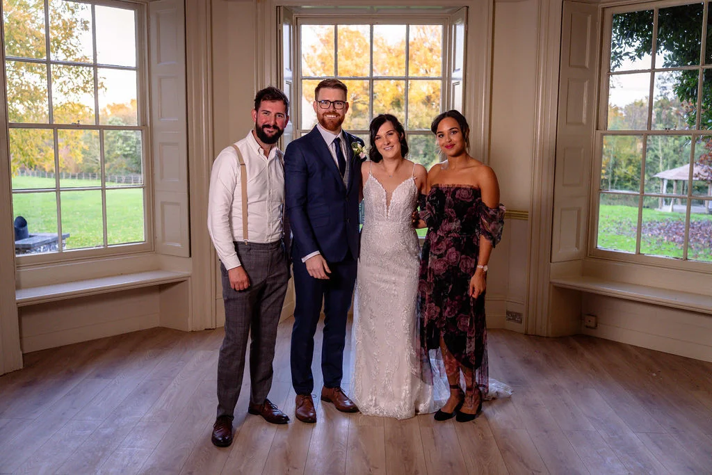 A newlywed couple posing indoors with two guests in a bright, wood-floored room featuring large Georgian sash windows overlooking autumn greenery.