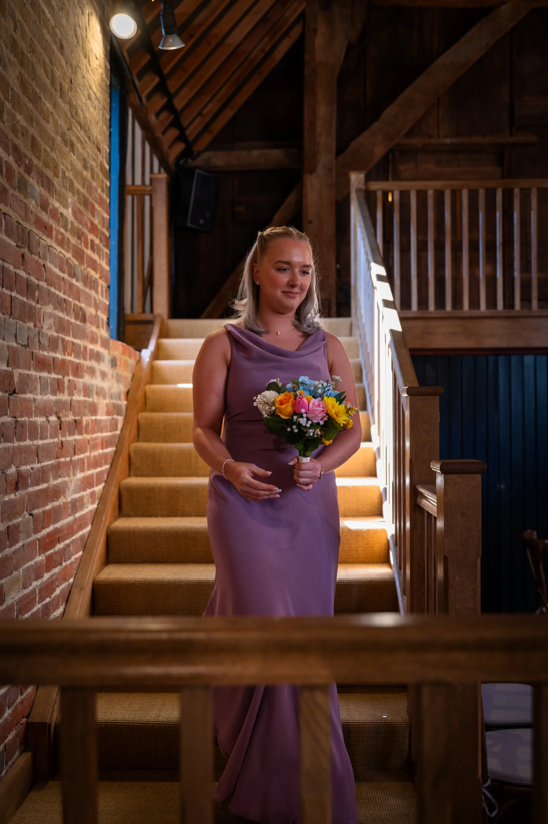 A bridesmaid in a mauve cowl-neck dress carrying a colourful bouquet descends a wooden staircase inside a rustic brick barn.