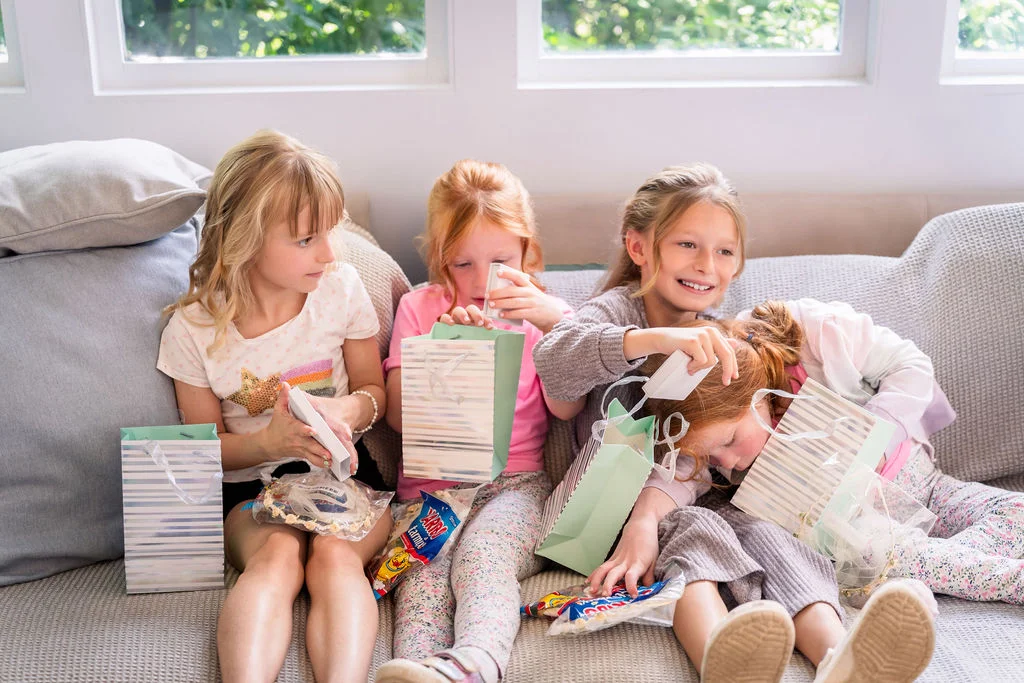 Four young flower girls happily sit together on a sofa opening gift bags and treats in bright, natural light.