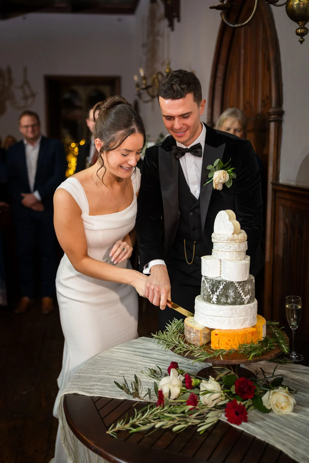 A smiling bride in a square-neckline white dress and a groom in a black tuxedo cut a multi-tiered cheese wheel wedding cake surrounded by white and red floral arrangements.
