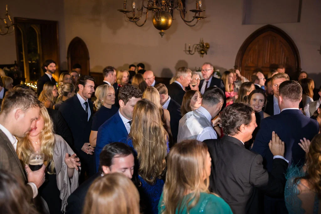 A bustling room of wedding guests in formal attire mingle during an evening reception under an ornate chandelier.