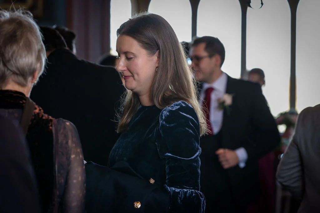 A wedding guest in a dark velvet dress mingles with other attendees indoors near tall windows.