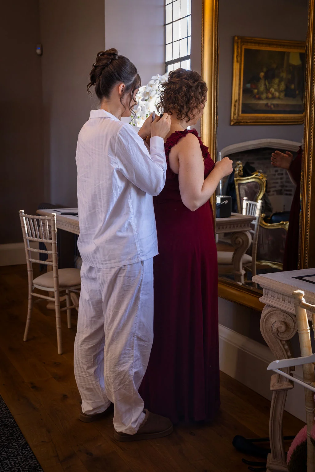 A bridesmaid in white pyjamas carefully buttons up a bride wearing a stunning red gown in front of a large ornate mirror.