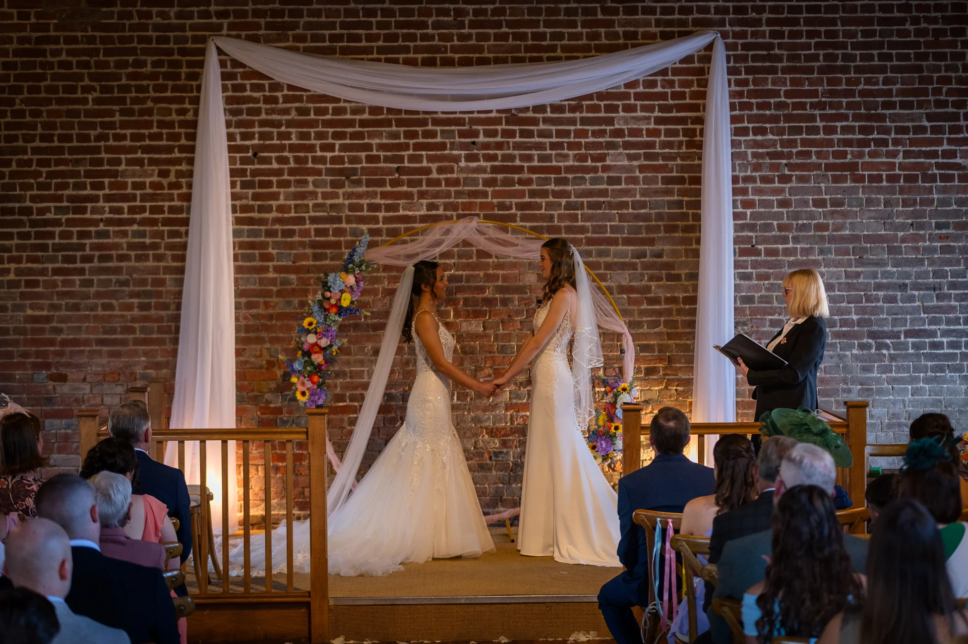 Two brides hold hands and exchange vows before a brick wall adorned with fabric draping and a floral circular arch during their indoor ceremony.