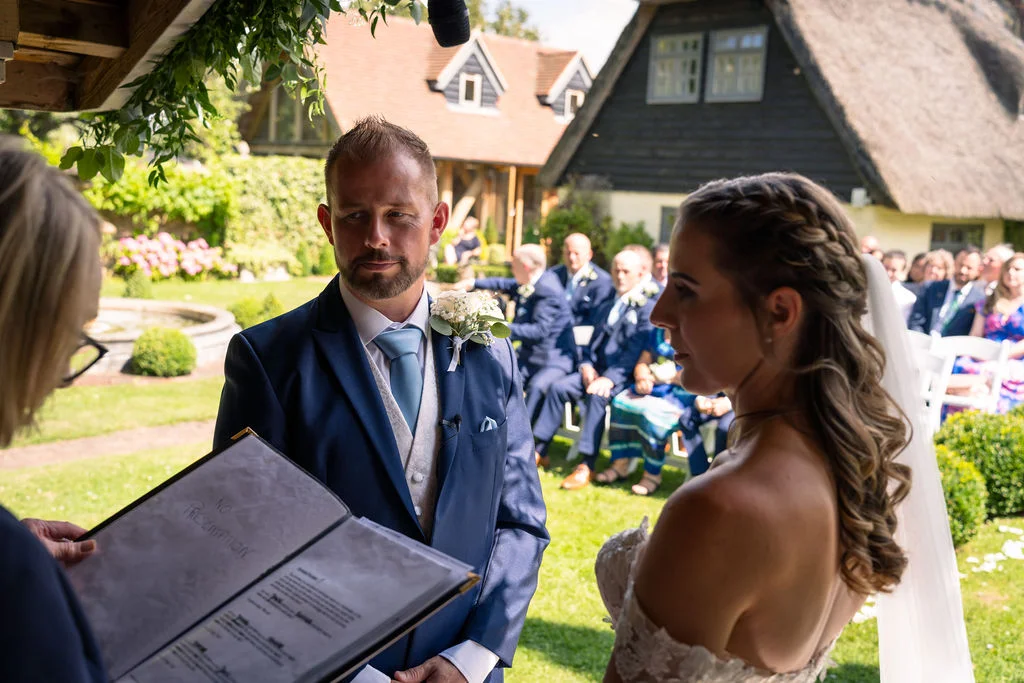 An outdoor wedding ceremony taking place on a sunlit lawn, with the bride and groom standing before a celebrant while guests watch from seated rows.