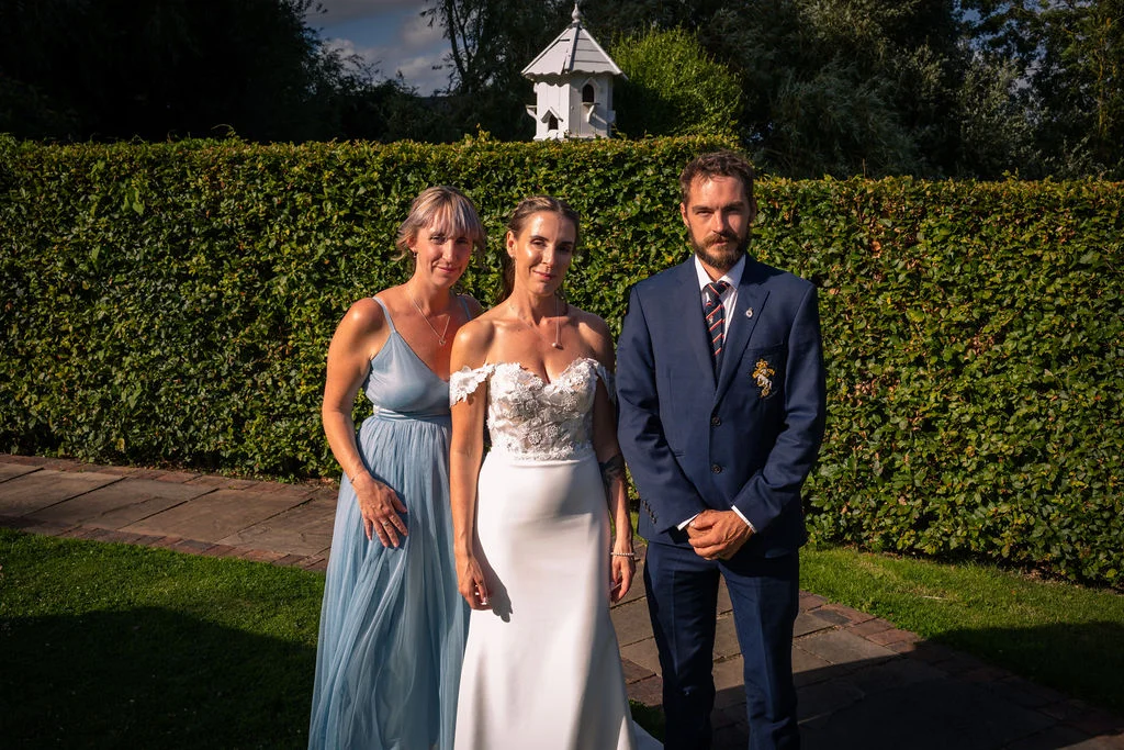 A bride in an off-the-shoulder gown, a groom in a navy suit, and a bridesmaid in a blue dress pose together outdoors in a manicured garden during golden hour.